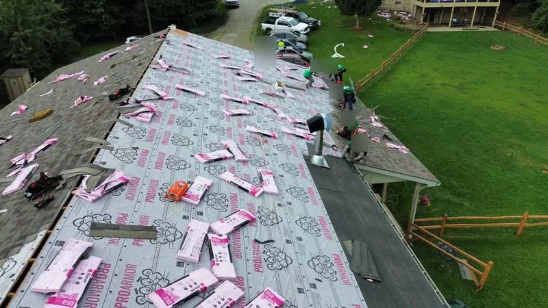 Roofing crew in green shirts shingling along the eave of a large roof covered in synthetic underlayment, with shingle bundles staged across it